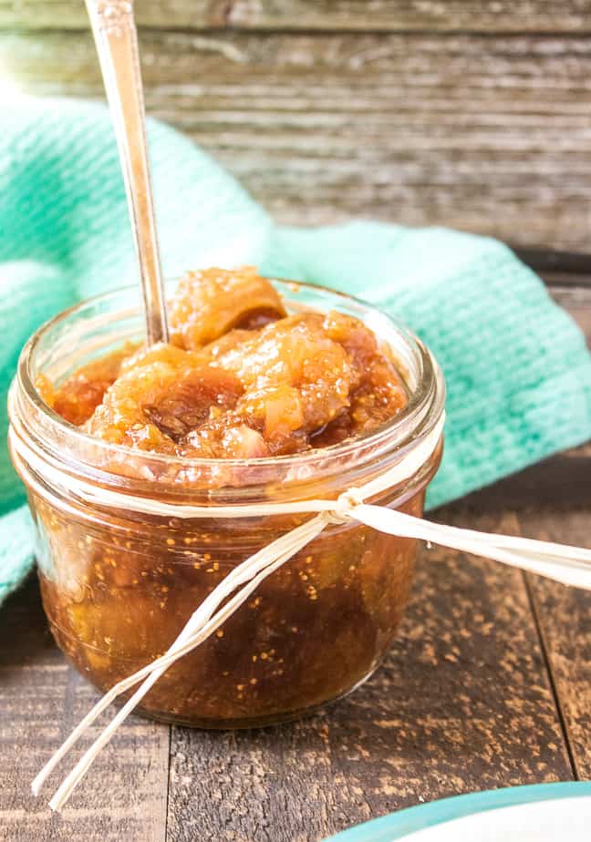 A small mason jar with a piece of raffia tied around the mouth, holds vanilla fig jam. There's a spoon in the jar, which sits on a wooden table. Behind the table is a mint green napkin.