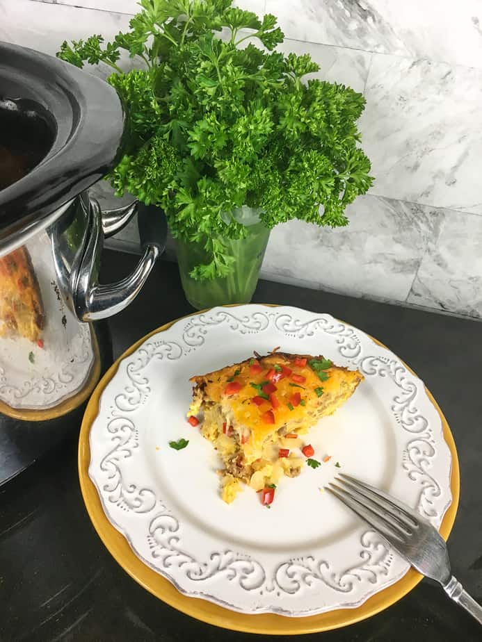 Slow Cooker Southwestern Breakfast Casserole wedge on a white plate with a fork. A vase with greenery in the background next to a slow cooker