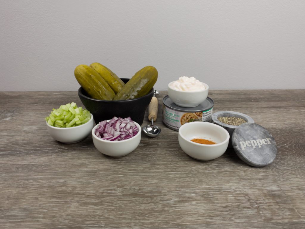 Ingredients for pickle tuna boats. chopped celery in a white bowl,chopped red onions in a white bowl, mayo in a white bowl, paprika and pepper in a white bowl, an unopened can of tuna and 3 pickles in a large black bowl. 