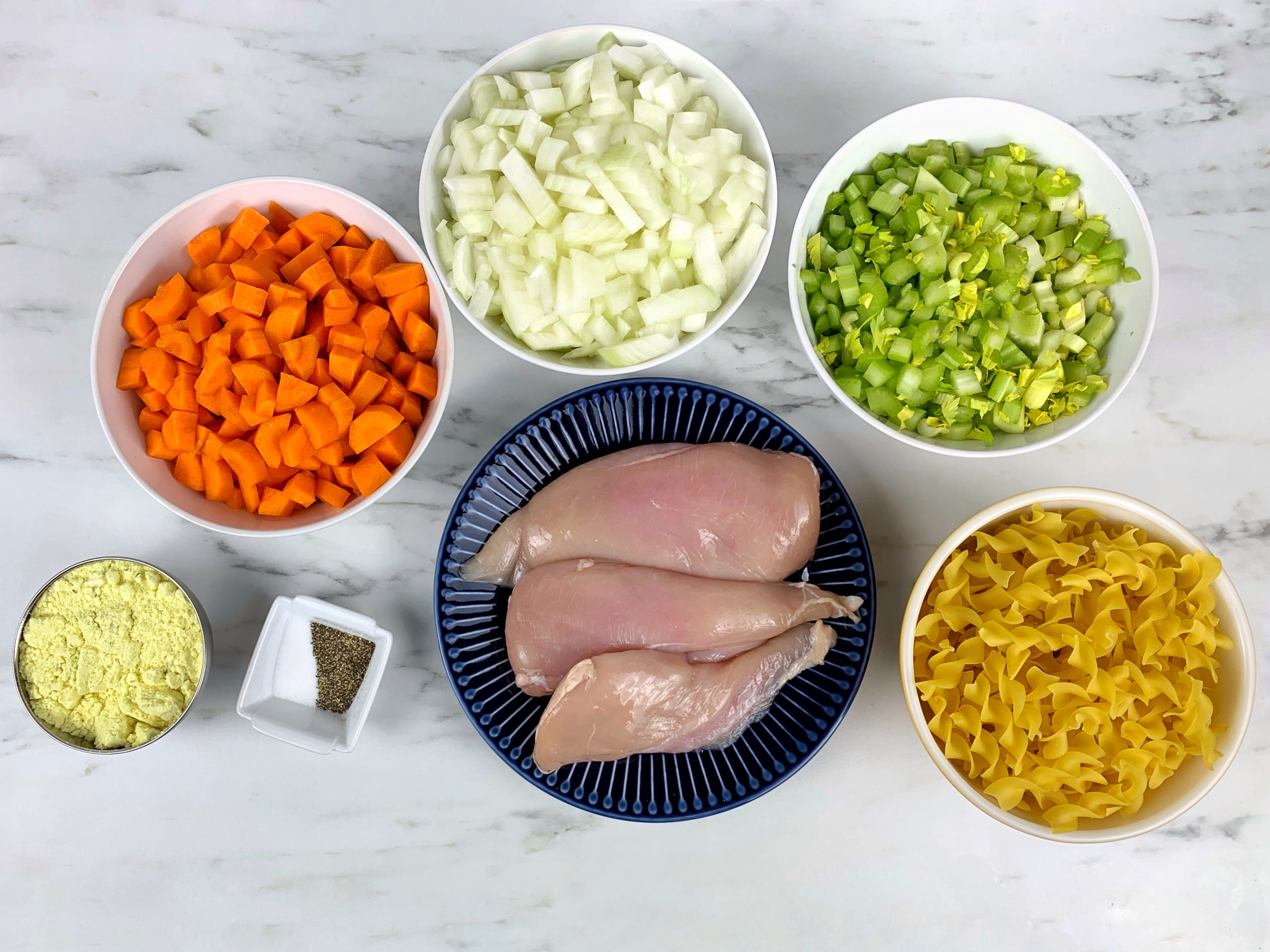 On a marble countertop, there's a black plate with raw chicken breasts in the center of the table. Around the chicken, forming a semi-circle are a small white bowl with pepper, a small black bowl with tarragon, a white bowl of carrots, a white bowl of onions, a white bowl of celery, a white bowl of uncooked egg noodles.