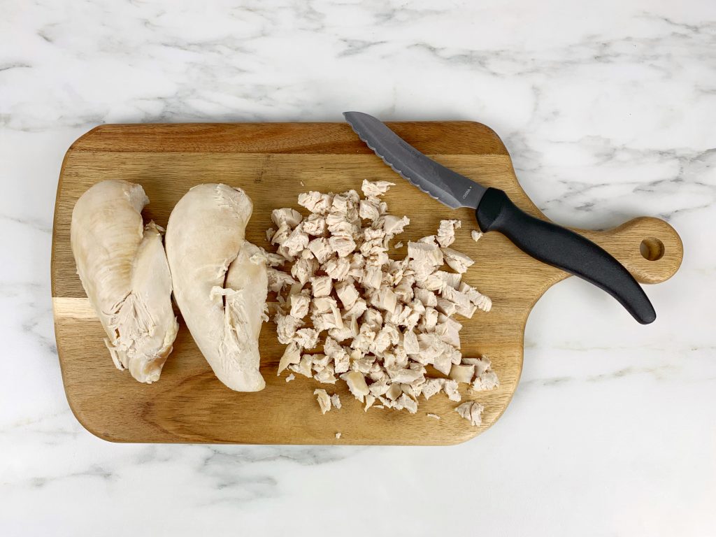 A wooden cutting board is on a white marble counter. There's two chicken breasts on the left-hand side of the cutting board. In the middle is cooked chicken, cut into chunks. On the right-hand side is a knife.