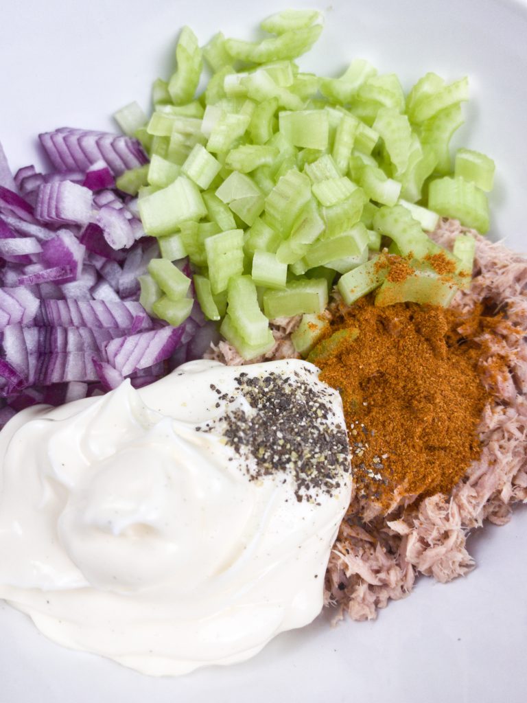 A plate with chopped onions and celery, mayo and tuna on it. spices are on top of the tuna and pepper is on the mayo.