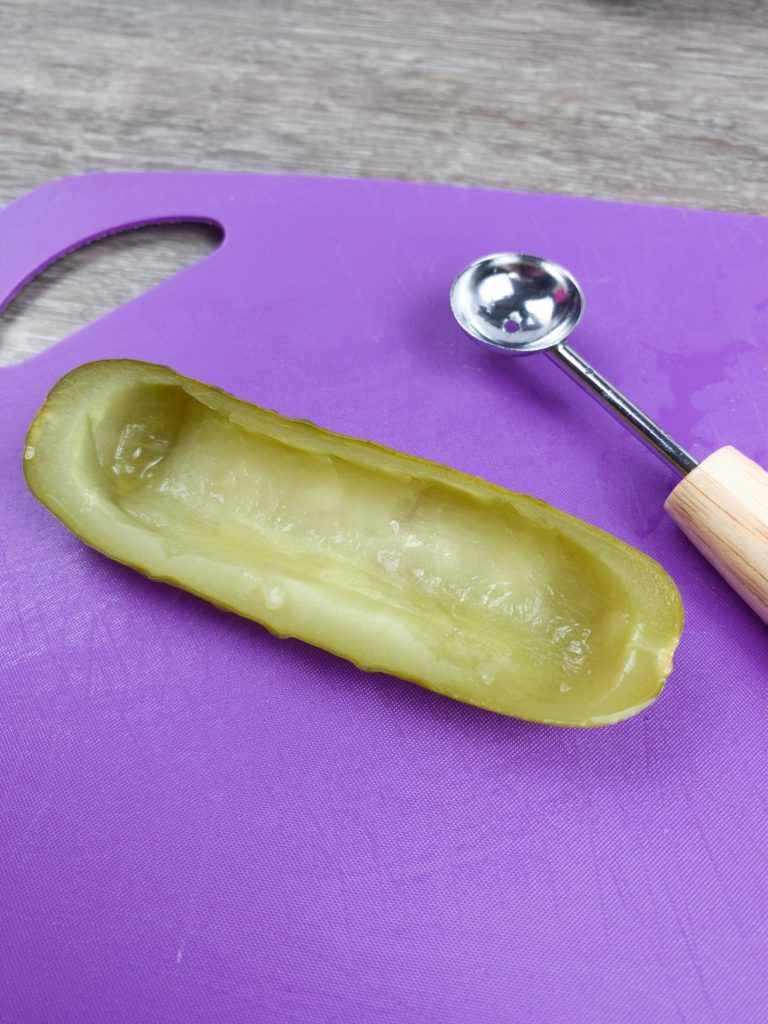 A pickle with the center hollowed out laying on a purple cutting board with a spoon next to it.