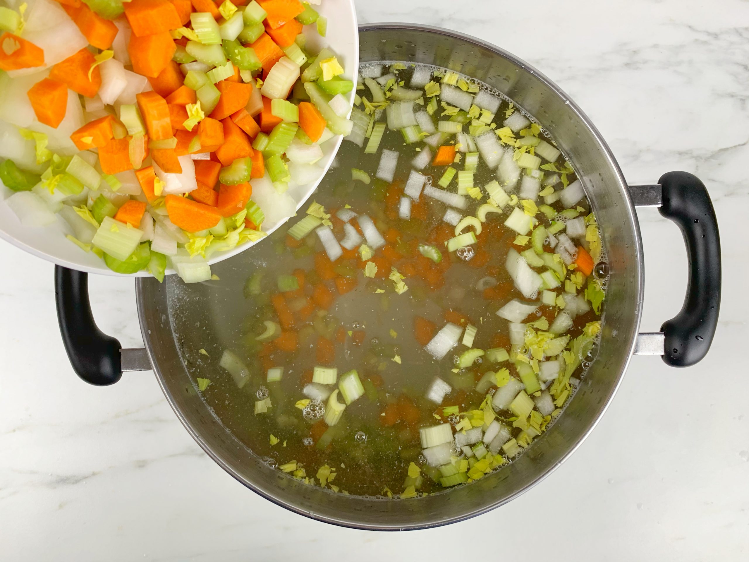 A pot of water on a marble countertop with celery, onions, and carrots in it. There's a white bowl above the pot with celery, onions, and carrots in it.
