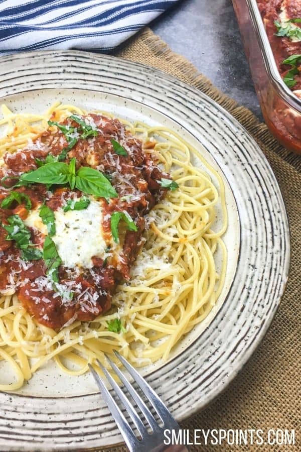 On a white plate, there's a bed of pasta. On the pasta site a piece of chicken parmesan with cheese and basil on top. The plate sits on a brown burlap place mat. 