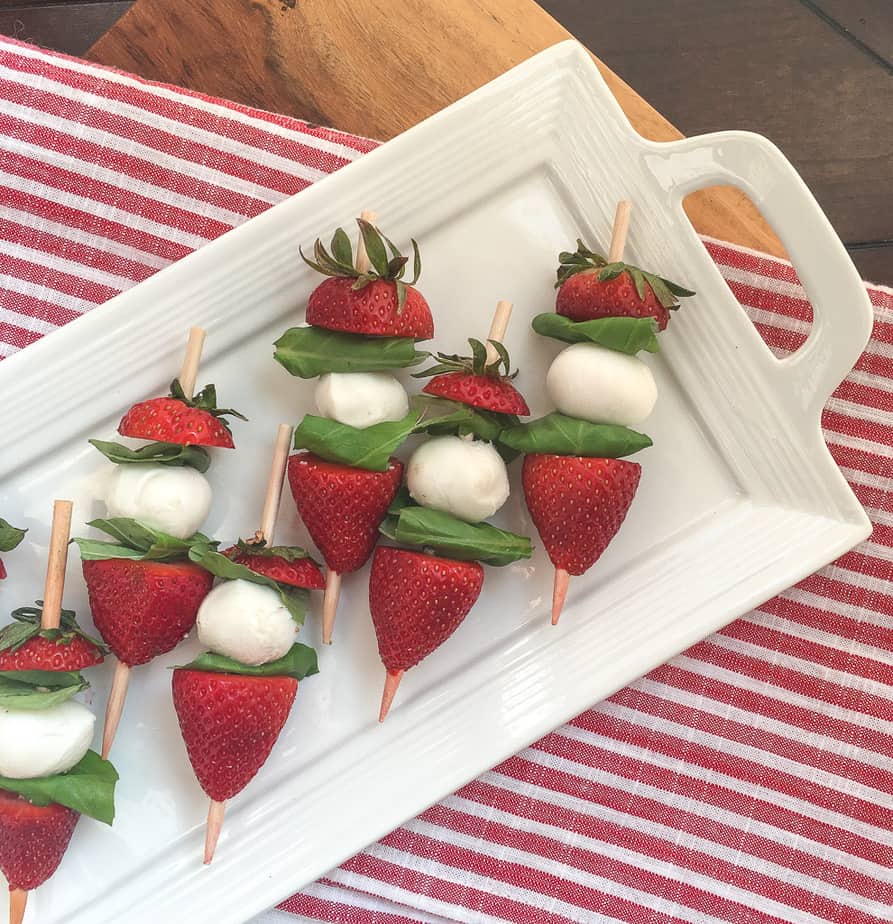 Seven skewers with strawberries, basil, and small balls of mozzarella cheese sit on a a white platter. The platter is on a red and white striped table cloth.