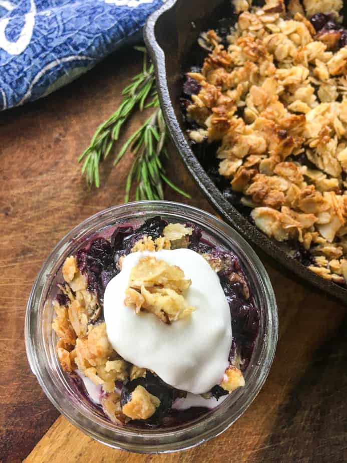 Blueberry crisp in a small mason jar with whipped cream and granola on top. The jar sits on a wooden table, next to a cast iron pan with more crisp in it. 