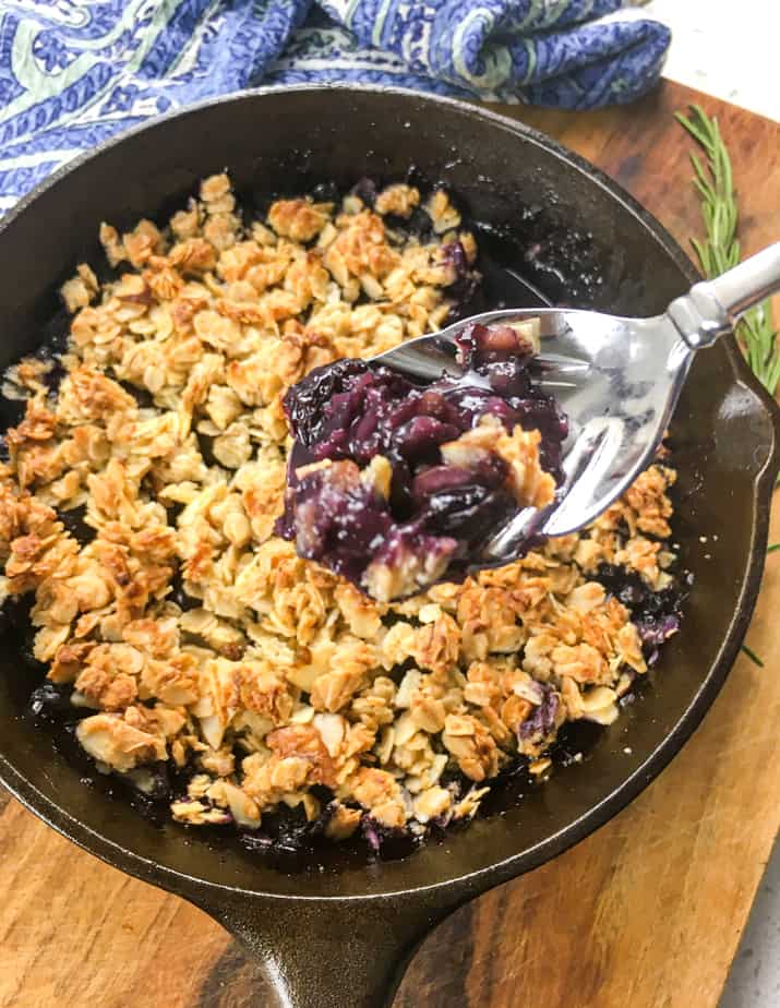 A cast iron pan with the crisp in it sits on a wooden table, with a sprig of rosemary next to the skillet. There's a spoon holding up a bit of the blueberry crisp, above the skillet. 