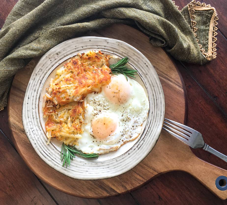 There's a wooden cutting board, with a grey cloth next to it. On the cutting board is a white plate with speckles on it. On t he plate are hash browns, sunny-side up eggs and a sprig of rosemary. There's an upside down fork on the board, touching the plate.
