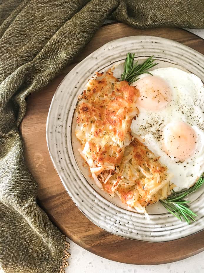 There's a wooden cutting board, with a grey cloth next to it. On the cutting board is a white plate with speckles on it. On t he plate are hash browns, sunny-side up eggs and a sprig of rosemary.
