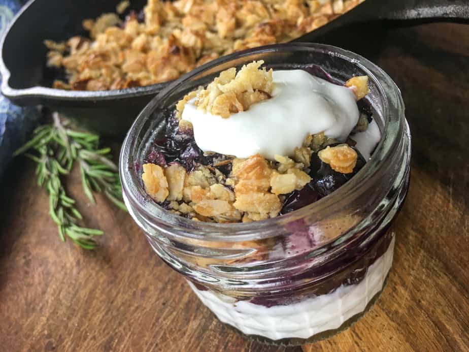 Blueberry crisp in a small mason jar with whipped cream and granola on top. The jar sits on a wooden table, next to a cast iron pan with more crisp in it. 