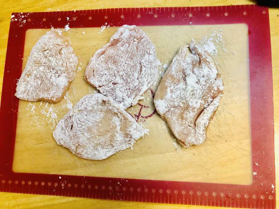 4 pieces of chicken breast, dredged in flour, on a cutting board that's clear with a red border. 