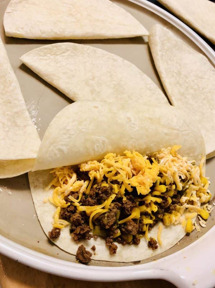 A huge round Pampered Chef stone with a white enamel lip. On top are tortillas folded in half, with one filled with cooked ground beef and cheese. 