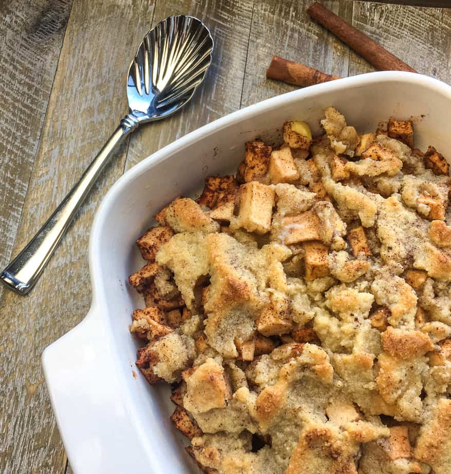 A white baking dish with handles on a wooden table with a spoon and cinnamon sticks next to it. The dish is filled with apples with a crust on top. 