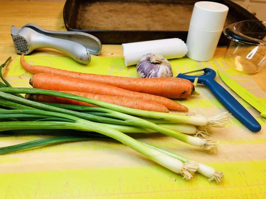 A green, flexible cutting board with a bunch of scallions, three carrots, a head of garlic with purple on it, a garlic press, a green vegetable peeler, a green paring knife, a measuring cup, a silicon garlic peeler, a small cylinder for slicing garlic, a large stone cookie sheet
