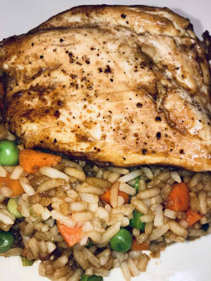 A piece of soy-glazed chicken on a bed of rice. The rice has chopped carrots and peas in it.
