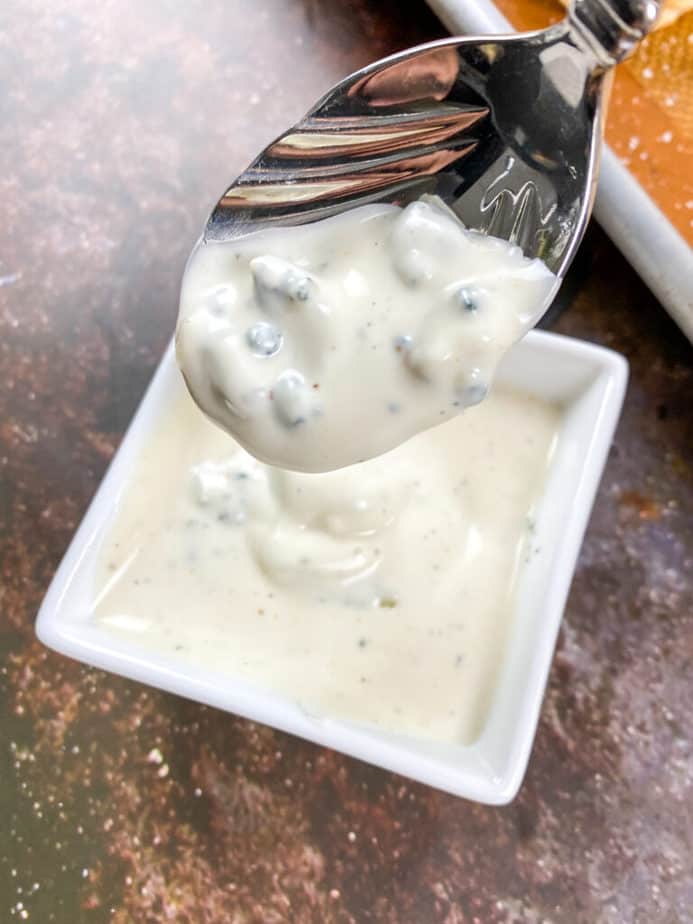 blue cheese sauce in a white, square ramekin. some sauce is in a spoon, which is held above the ramekin.
