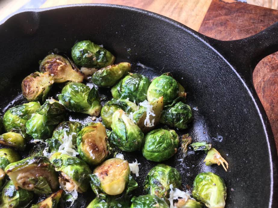 A cast-iron pan, filled with lightly browned Brussels sprouts lightly dusted with shredded cheese. The pan is on a wooden table.