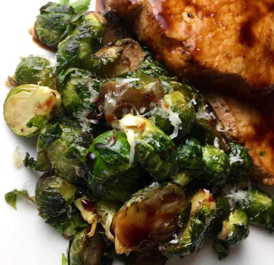 A white plate with a pile of Brussels sprouts with some maple balsamic glaze and a sprinkling of cheese on them. On the plate, in the upper right corner, there are boneless pork chops.