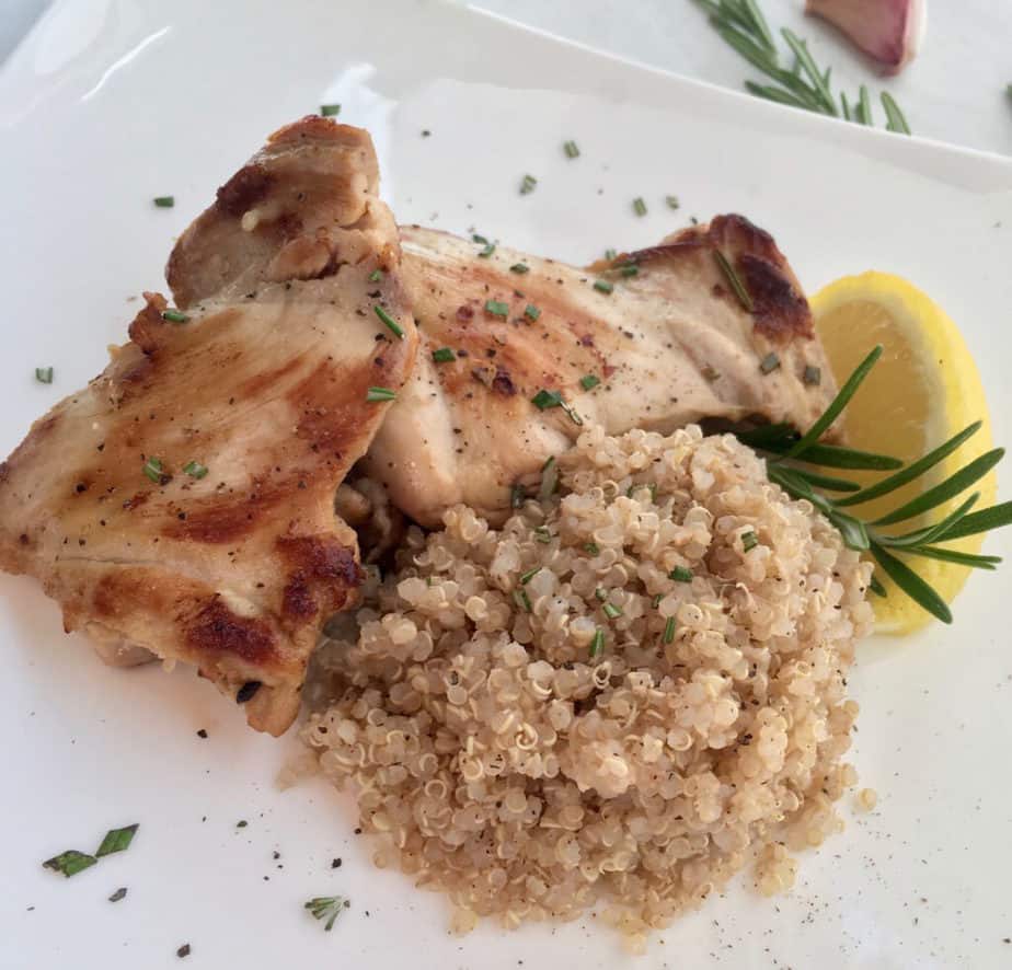 A nicely browned chicken thigh sits on a white plate. To the right is a pile of rice, on top of the rice and chicken is a sprig of rosemary and a lemon wedge.
