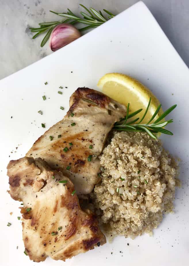 A nicely browned chicken thigh sits on a white plate. To the right is a pile of rice, on top of the rice and chicken is a sprig of rosemary and a lemon wedge.