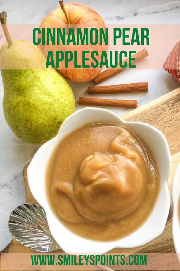 There's an apple on the top center, with 2 cinnamon sticks under it. TO the left of that is a pear. These items are on a marble counter top. There's a wooden cutting board with a small white cup on it. In the cup is applesauce. There's a spoon next to the bowl. 