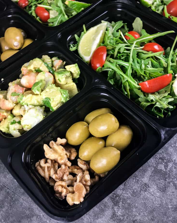 A bento box with field greens, sliced grape tomatoes and a lime wedge in one square, the keto shrimp avocado in one square, green olives and walnuts in a square, green olives in another square