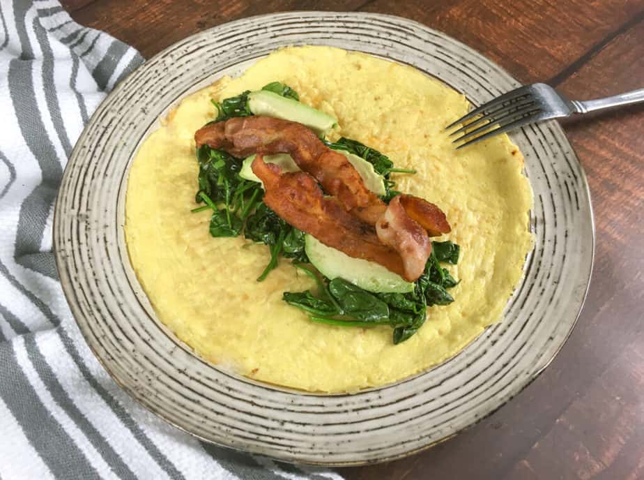 An off-white plate sits on a wooden table. There's a white napkin with grey stripes on the upper left, under part of the plate. On the plate is an open-faced egg wrap. In the middle is a pile of spinach, topped with avocado and then bacon. There's an upside down fork on the edge of the plate. 