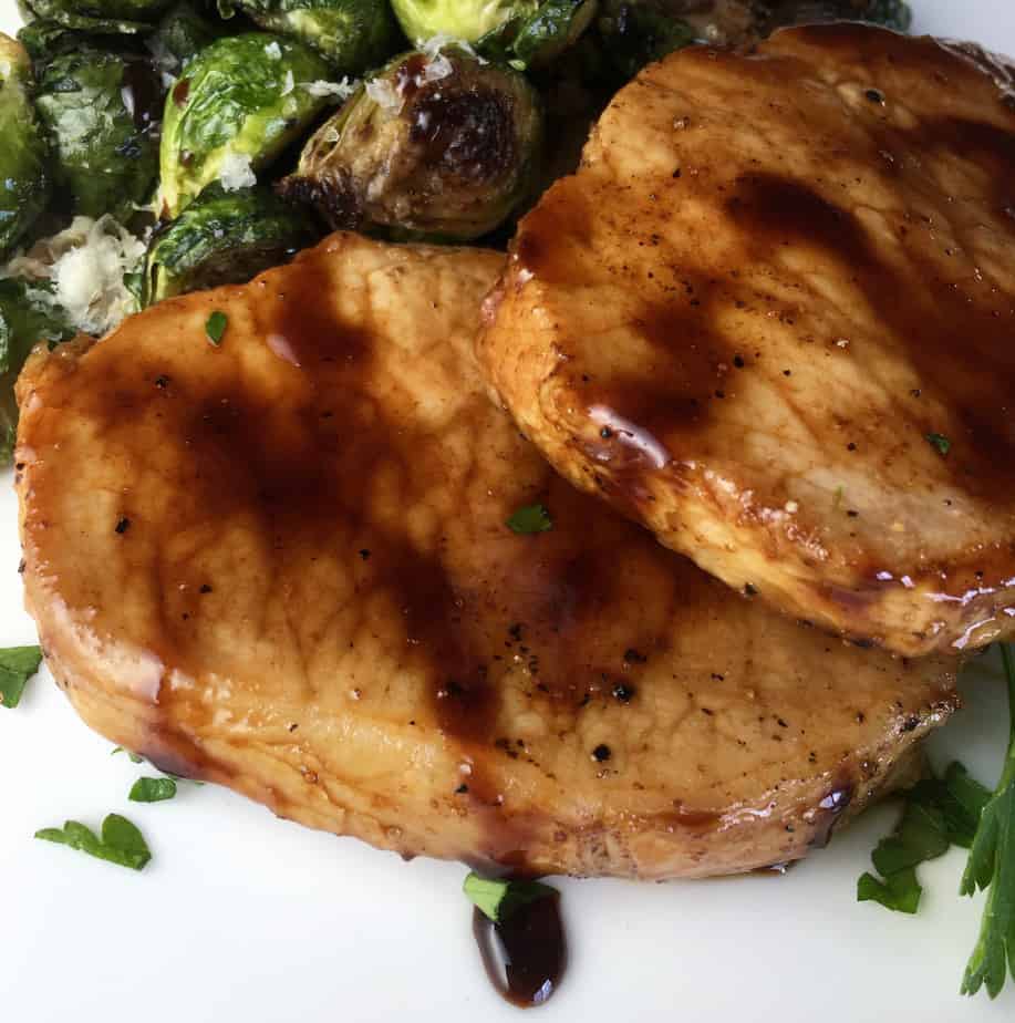 Two pieces of pork, on a white plate, with sauce on them and roasted Brussels sprouts on the top left of the plate.