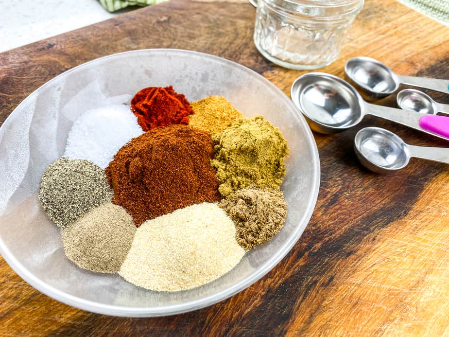 A variety of spices in a bowl and measuring spoons to the left of the bowl. 
