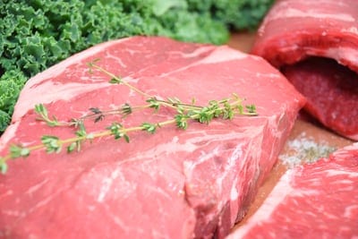 A boneless pork chop on a wooden cutting board with a sprig of thyme on it. There's a bunch of parsley in the background.
