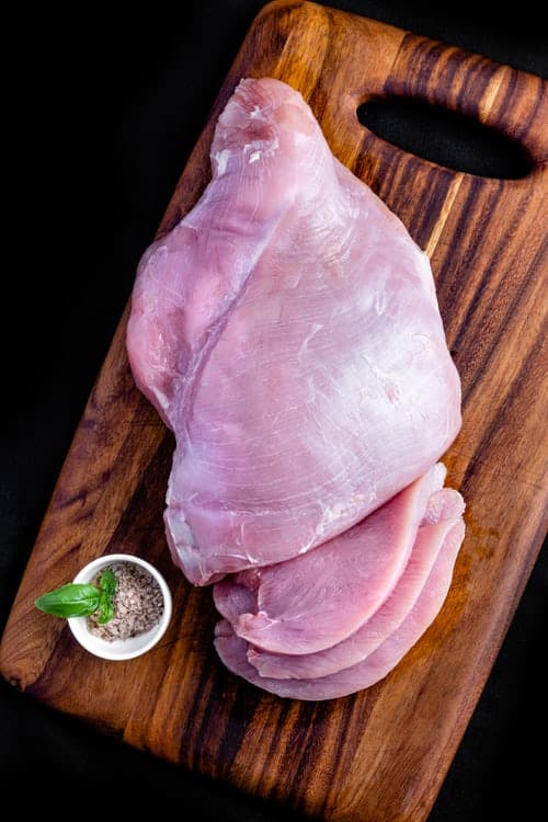 A wooden cutting board with a chicken breast on it. It's mostly a whole breast, with a few slices cut on the end. There's a small white cup with some herbs in it and a basil leaf sticking out of it.
