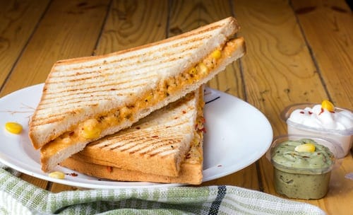 A white plate sits on a wooden table. On the plate is a grilled cheese sandwich, cut diagonally. It has grill marks on it. There's a small cup of a green dip next to the plate and a bigger cup of a white dip to the top right of that cup. 