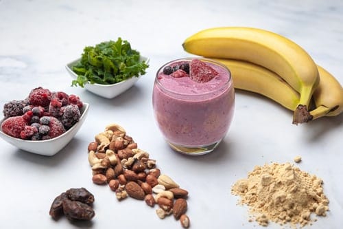 On a white table there's a small glass with a purple smoothie in it. There are a few pieces of fruit on the top of the glass. On the table, in a circle around the glass are bananas, greens, berries, nuts and protein powder. 