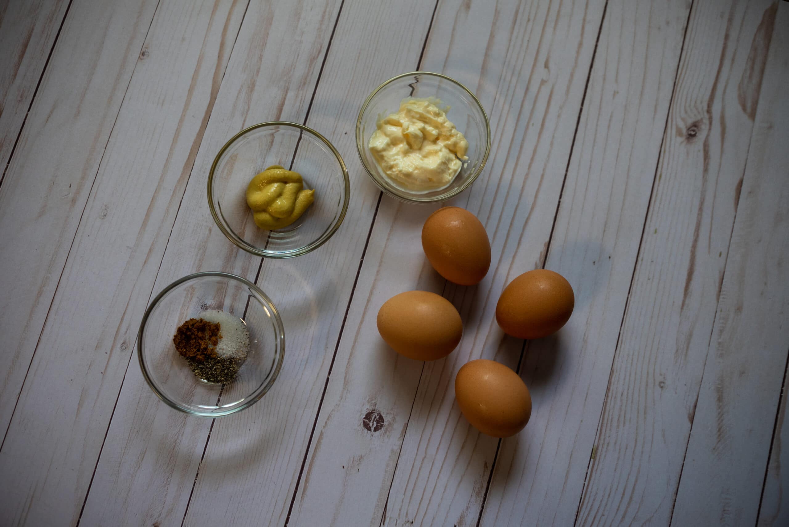 Ingredients for curried deviled eggs. Brown eggs and ingredients in clear bowl on a wood table 