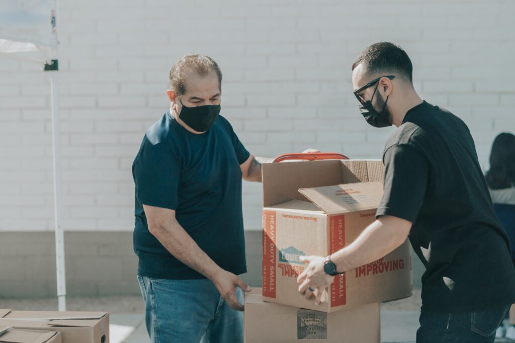 volunteer together. Two men lifting a box both wearing masks.
