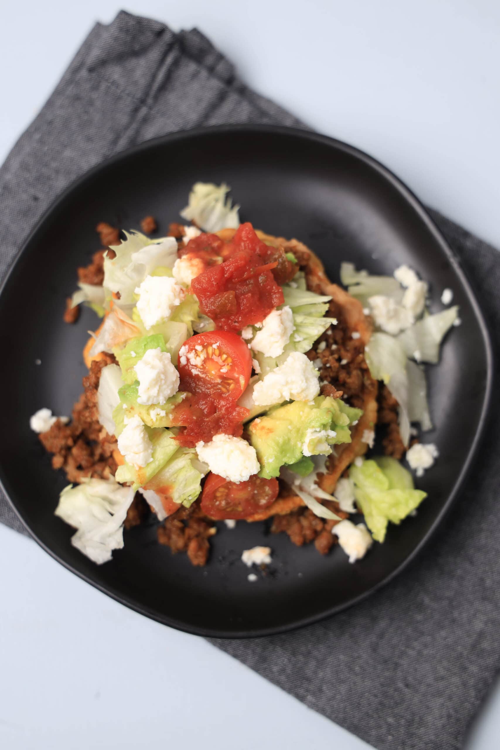 A sope on a black plate with ground beef, tomatoes, cheese, and avocado toppings