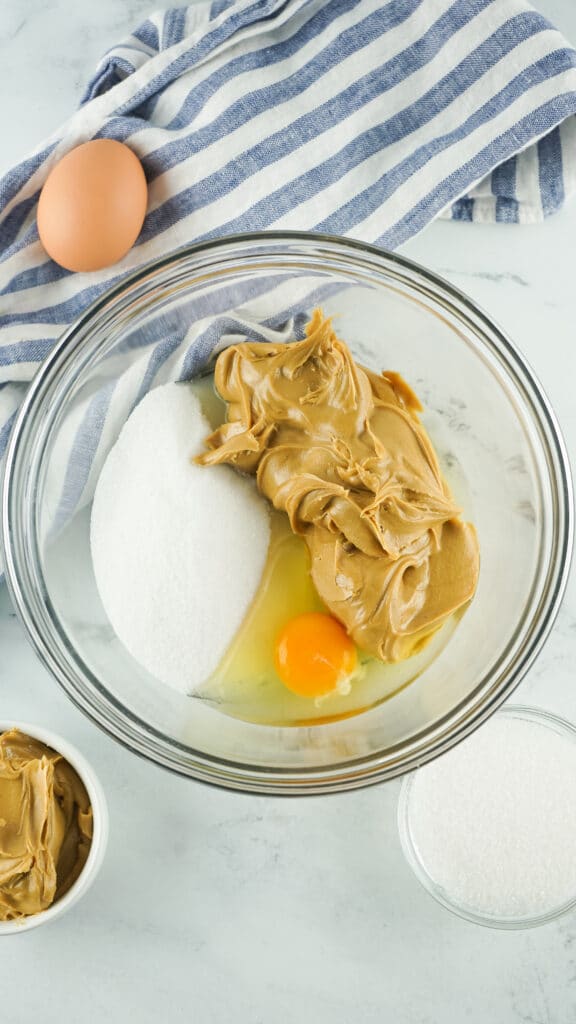 A glass bowl with peanut butter, sugar and an egg in it. a small cup of peanut butter and sugar are on the sides of the bowl. a blue and white dish towel is in the background