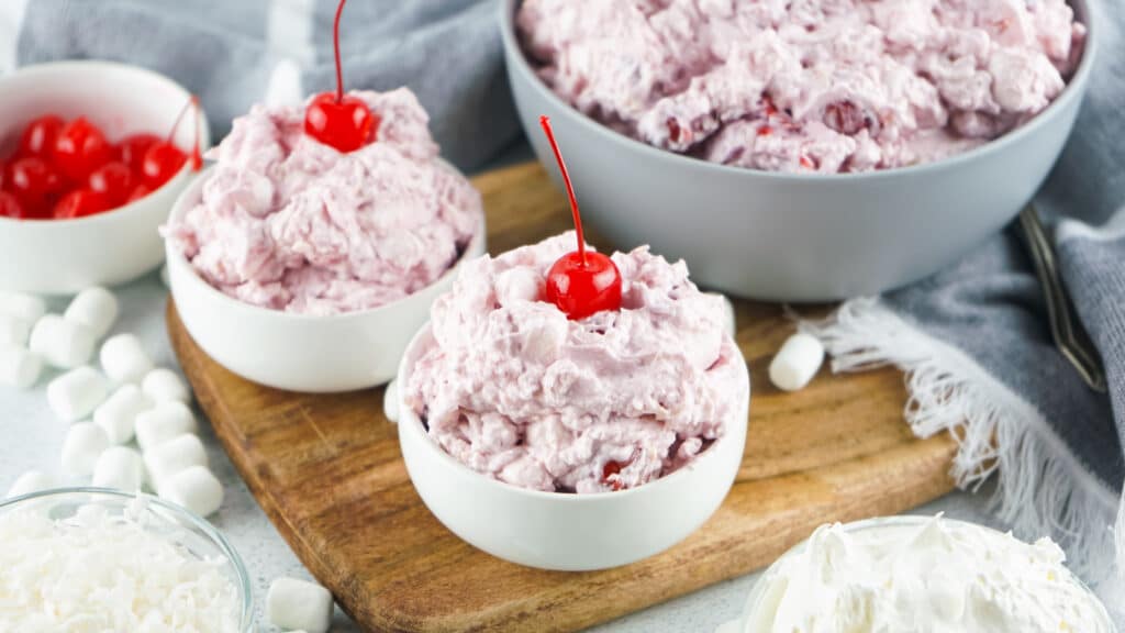 cherry fluff in 2 small white bowls and a larger portion in a grey bowl in the back. the bowls are on a wooden cutting board. There is a grey towel and a spoon on the right side.