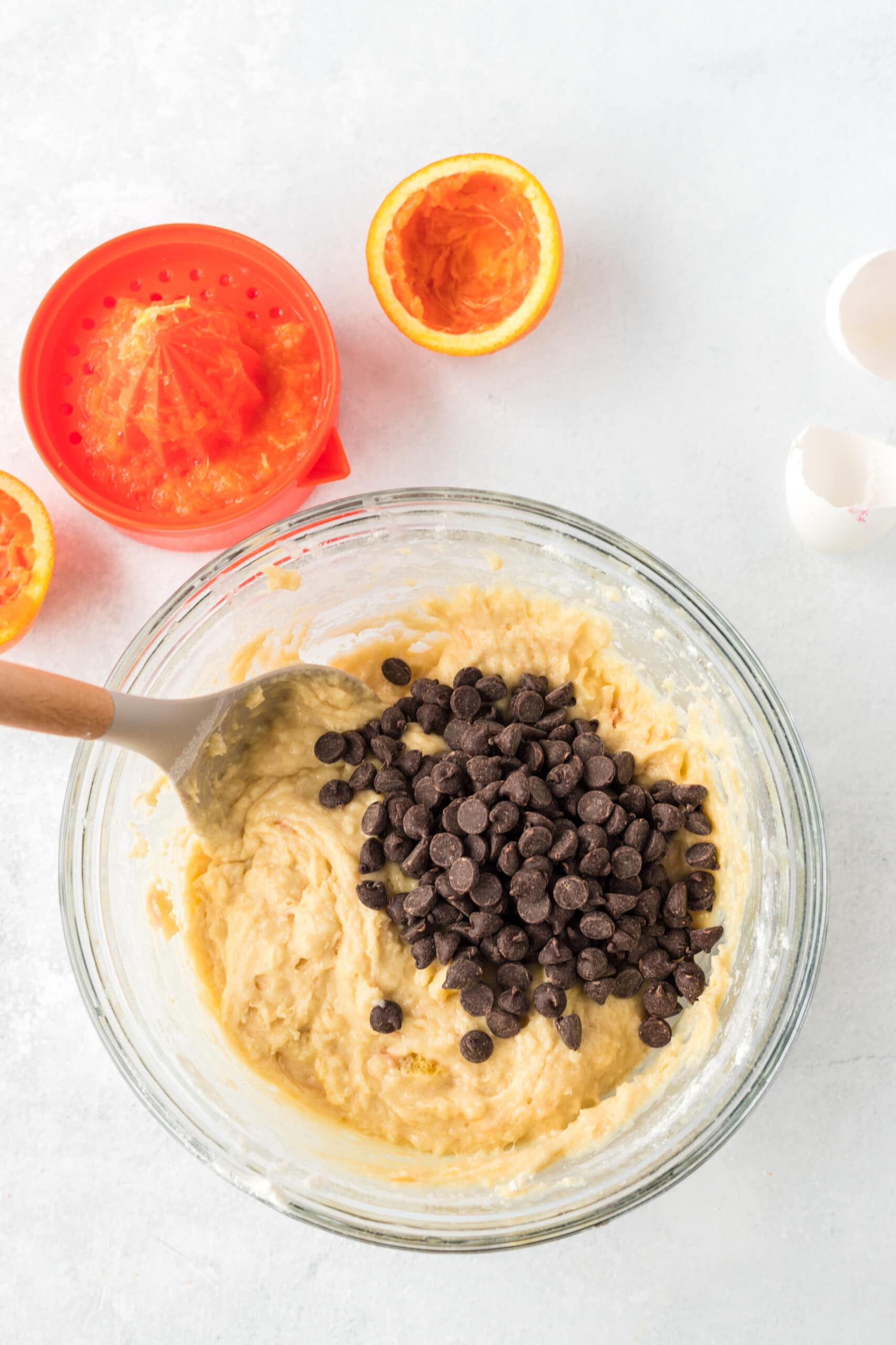 An orange that has been juiced next to a hand juicer and a clear bowl with chocolate chips and batter in it, being mixed with a wooden spoon