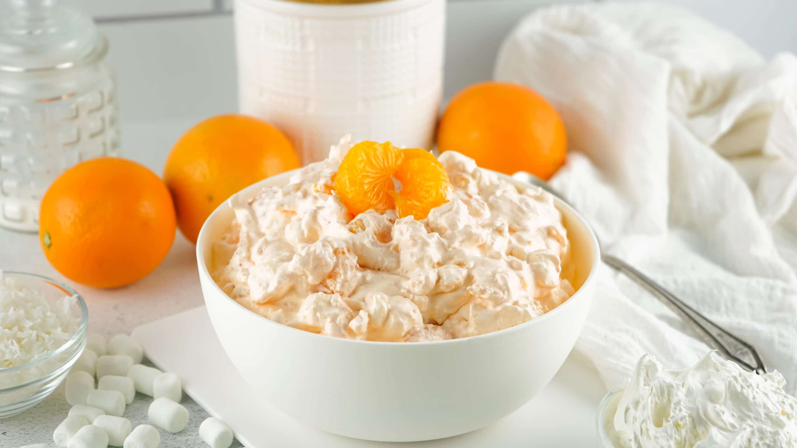 Orange fluff mixed in a white bowl. Oranges and a bowl of shredded coconut in the background 