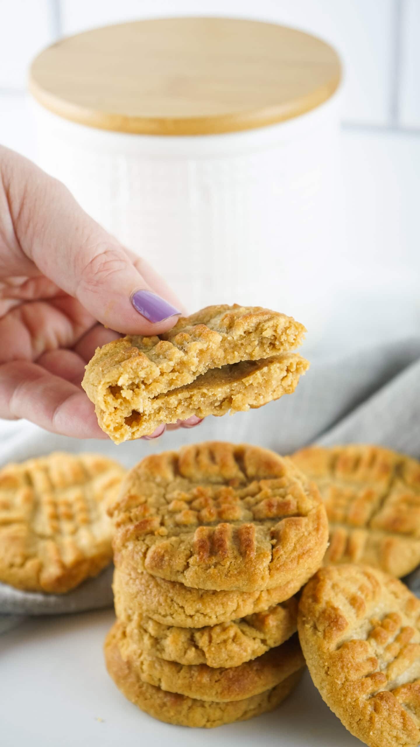 A hand holding a peanut butter cookie that has been broken in half. several peanut butter cookies are stacked and arranged on a white table 
