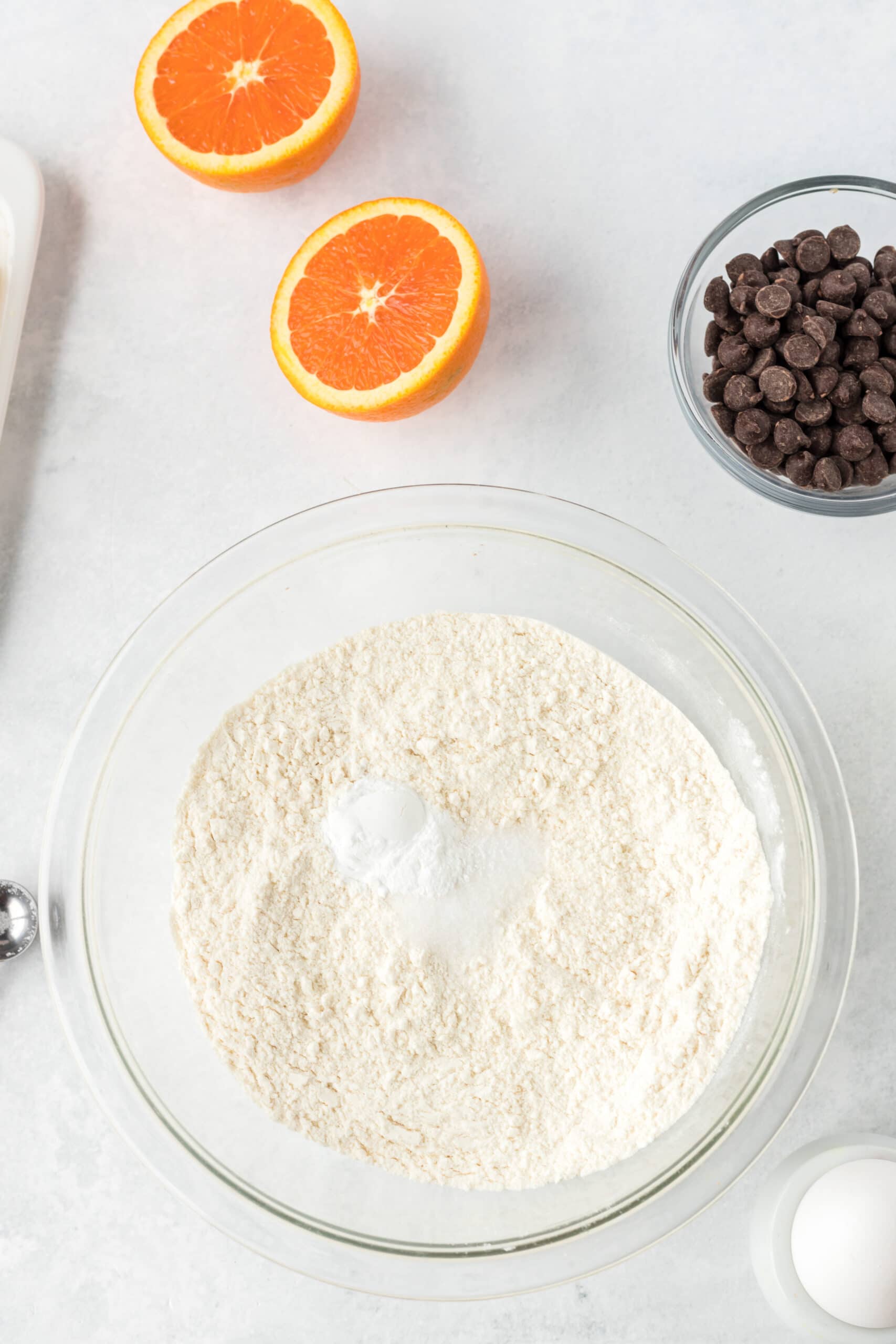 an orange cut in half and a bowl of flour and chocolate chips in clear bowls on a white table.