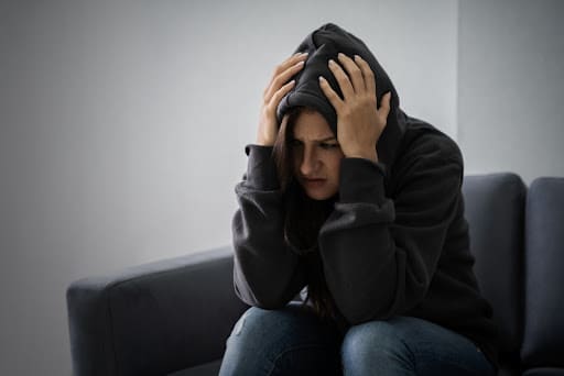 Young Depressed Woman In Hoodie Sitting On Sofa At Home
