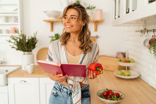 Funny housewife woman looking at recipe in cookery book preparing vegetable salad cooking food in light kitchen at home. Dieting healthy lifestyle concept.