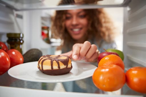 Woman reaching for chocolate donut inside refrigerator stocked with fruits and vegetables.