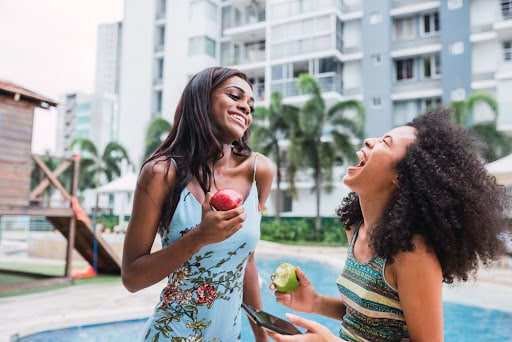 Cheerful ethnic women laughing while eating apples at poolside resort.
