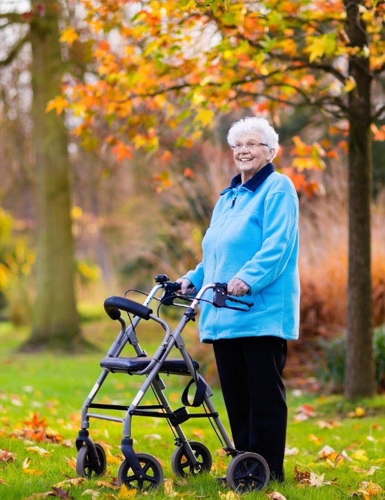 An elderly woman with an assistive device walker is seen smiling in the park during autumn.