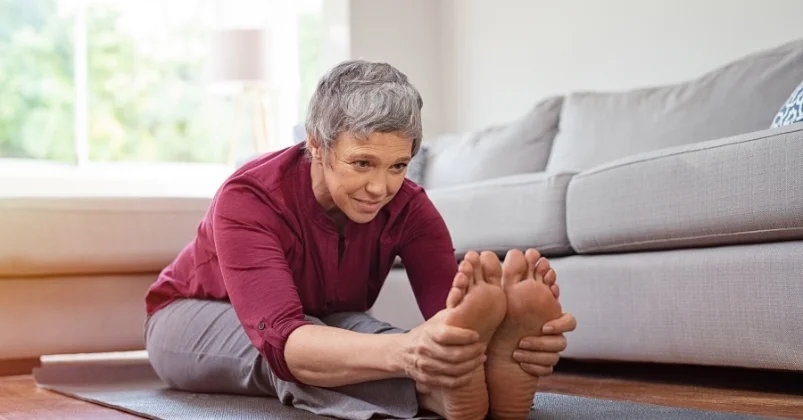 In an indoor setting elderly woman is sitting on the floor engaging in a stretching exercise. With remarkable flexibility she bends forward and holds her feet with her hands while sitting on a mat