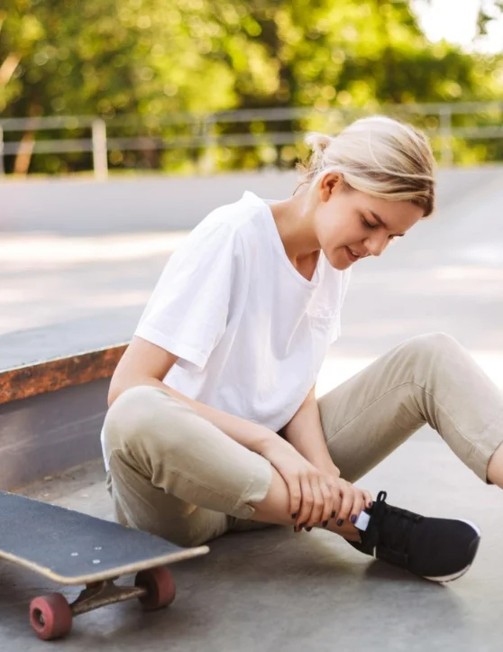 In her white top and light brown pants, a young skater girl is seen holding her painful leg while clutching onto her skateboard